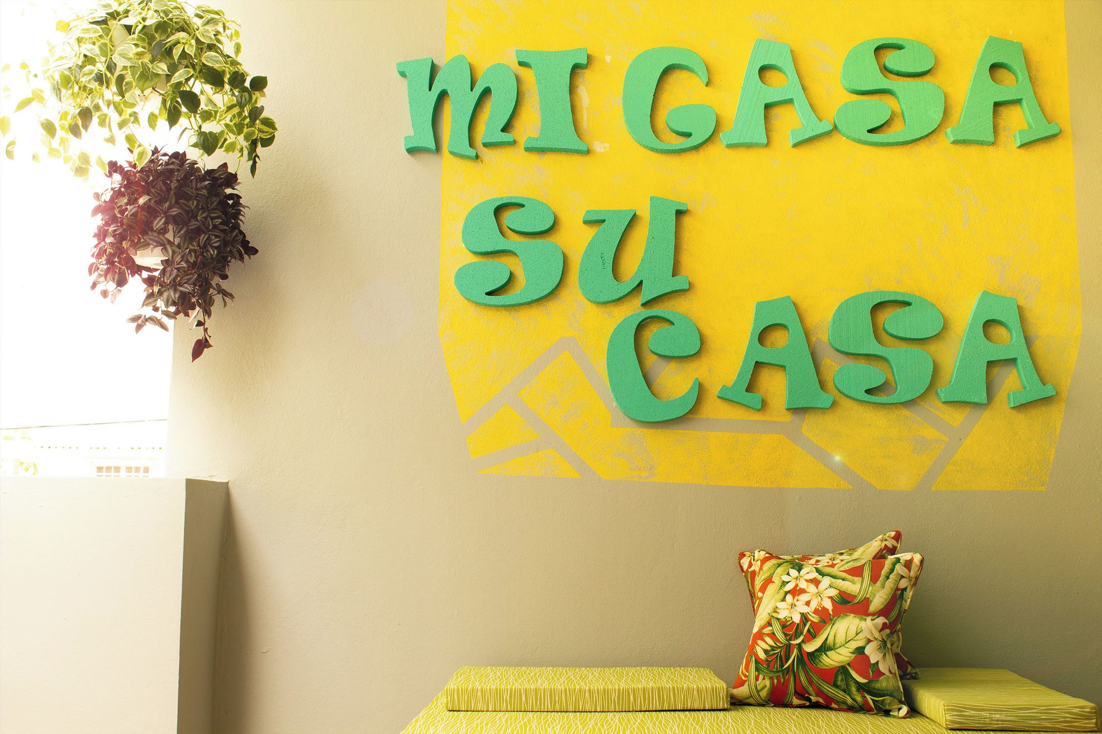 A room with a yellow wall, a green sign that reads "MI CASA SU CASA", a hanging plant, a patterned pillow, and a small yellow bench.
