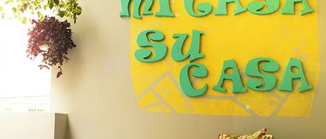 A room with a yellow wall, a green sign that reads "MI CASA SU CASA", a hanging plant, a patterned pillow, and a small yellow bench.
