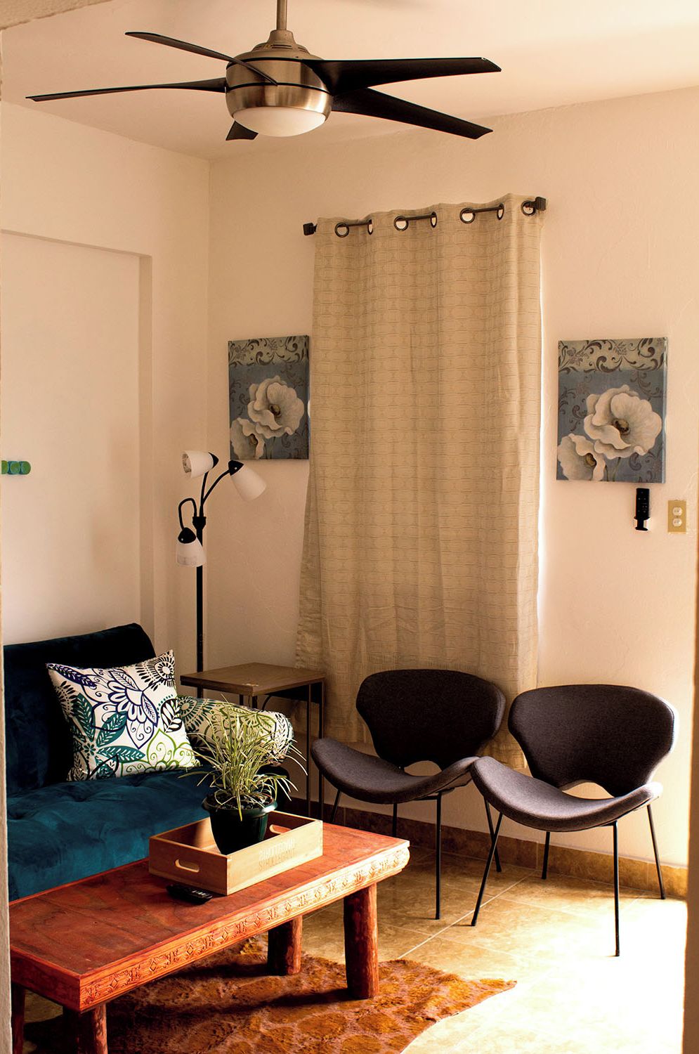Ein Wohnzimmer mit Deckenventilator, zwei Sessel, einem kleinen Tisch mit Pflanze, einer Couch mit gemusterten Kissen und zwei Wandbildern.