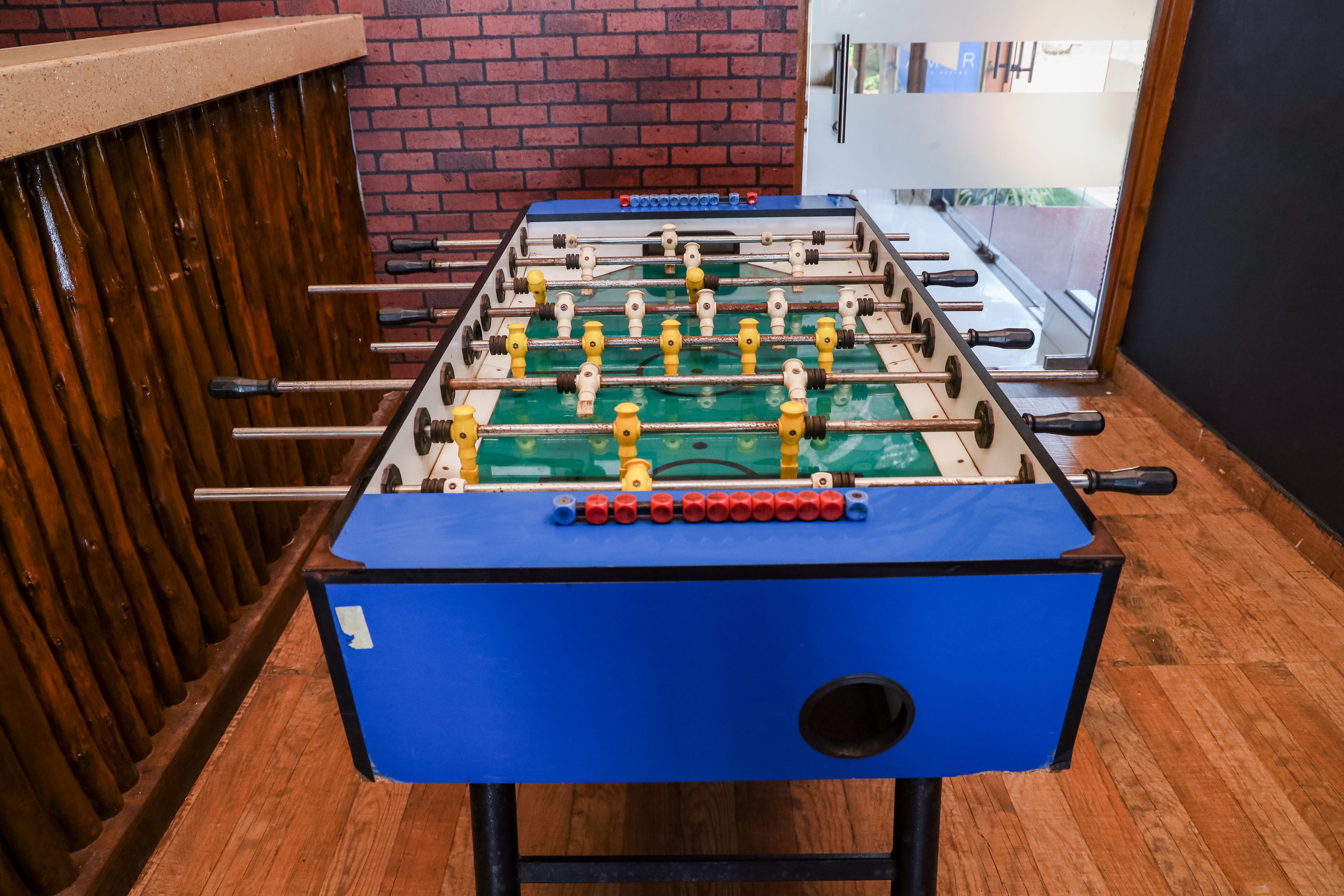 Una mesa de fútbol de mesa con superficie azul y figuras de jugadores amarillas y rojas, frente a un muro de ladrillo y suelo de madera.