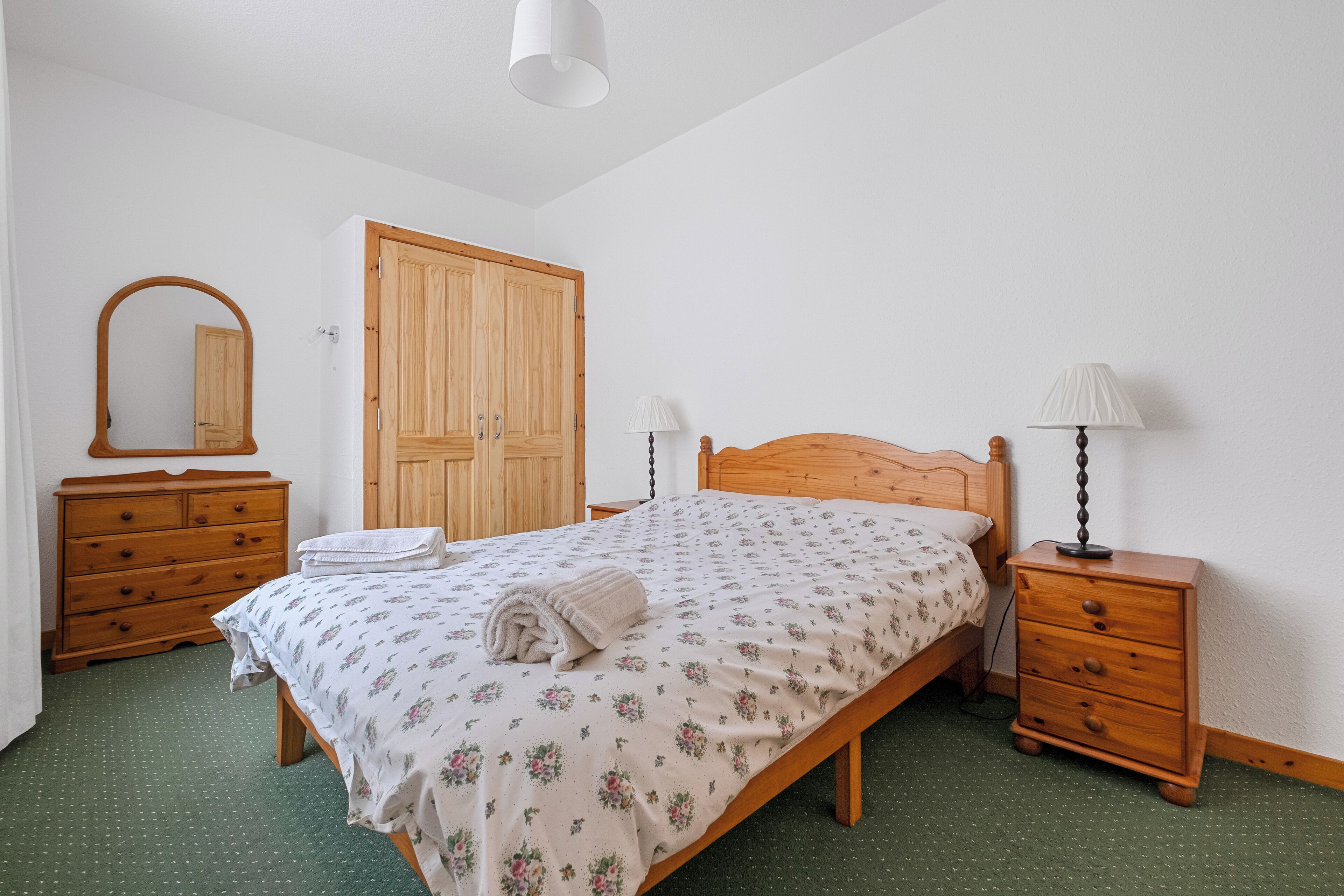 Een slaapkamer met een houten bed, een nachtkastje, een dressoir, een kledingkast en een spiegel.