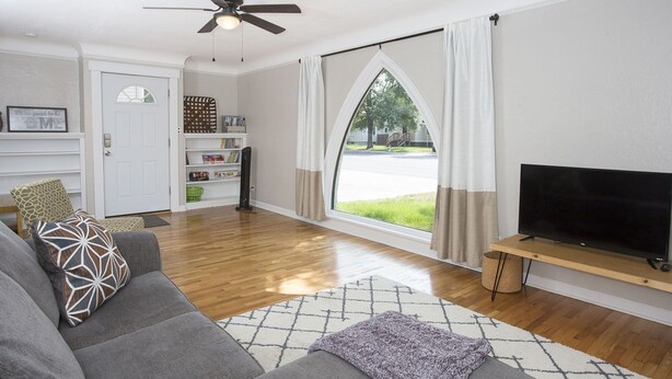 A living room with a grey sofa, a television on a wooden stand, a ceiling fan, and a large window with curtains. - Triangular Casa in Billings (Billings)