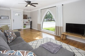 A living room with a grey sofa, a television on a wooden stand, a ceiling fan, and a large window with curtains. - Triangular Casa in Billings (Billings)