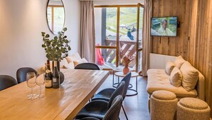 Ein modernes Wohnzimmer mit Holzboden, einem Esstisch mit Stühlen, einer Couch und einem an der Wand befestigten Fernseher.