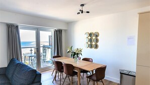 Un salon moderne avec un canapé, une table à manger et des chaises. Il y a un balcon avec vue sur la mer et les bâtiments.