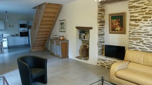 Un salon avec un canapé, un téléviseur, un mur en pierre, une cheminée et une cuisine à l’arrière-plan.