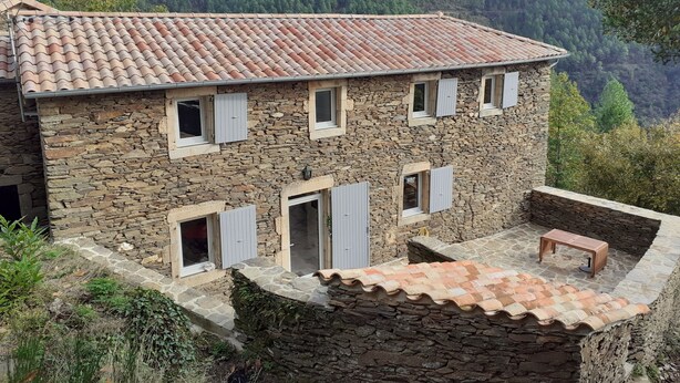 Un bâtiment en pierre avec un toit en tuiles, de nombreuses fenêtres dotées de volets et un petit espace extérieur aménagé pour s’asseoir.