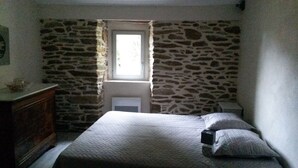 Une chambre avec un mur en pierre, une fenêtre, un lit avec des oreillers et une commode.