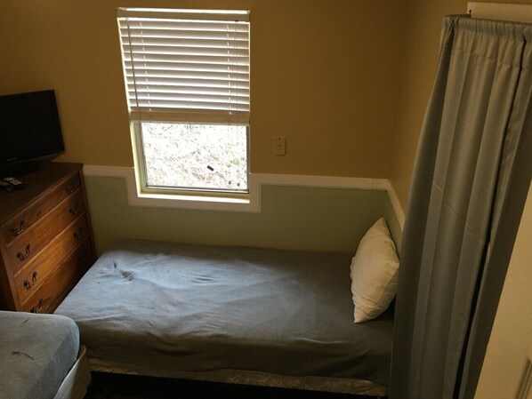 A small, single-bed room with a window, a wooden dresser, and a television.