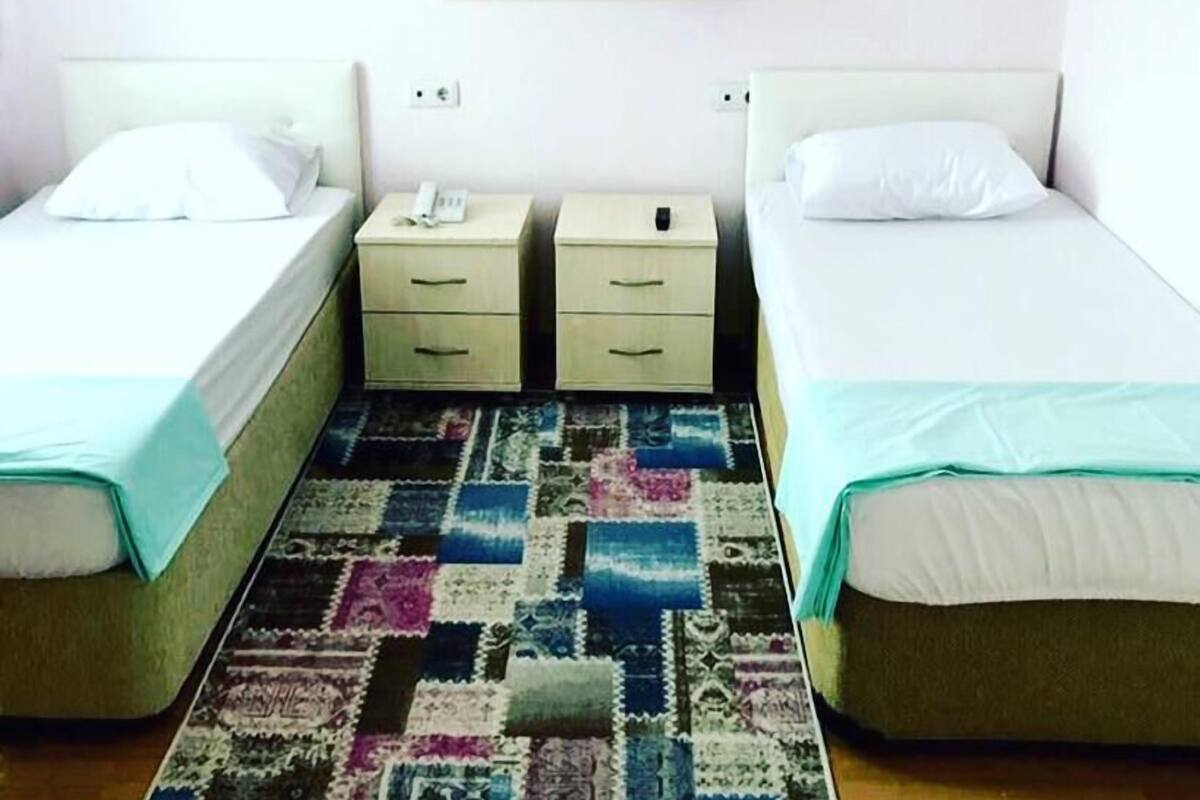Una habitación con dos camas individuales, una mesita de noche con teléfono, estante en la pared y una alfombra colorida.