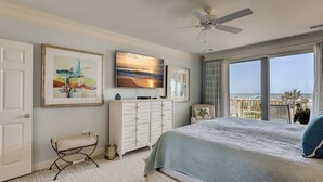 Un dormitorio con cama, cómoda, silla, ventilador de techo, televisor y balcón con vista al océano.