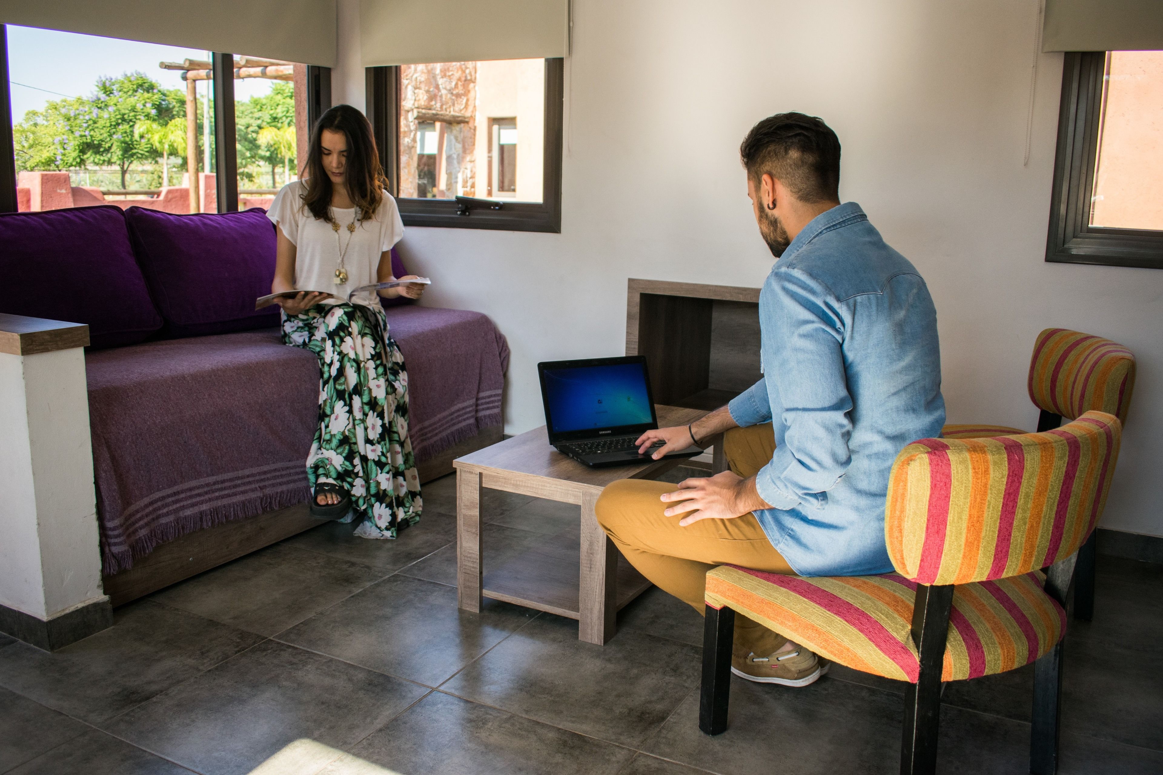 Een kamer met een bank, een stoel, een laptop op een bijzettafeltje, en een raam met uitzicht op groen.