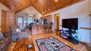 A spacious living area with a high ceiling, wooden flooring, a central island with bar stools, a flat-screen TV, and a cozy seating area with a patterned rug. - 2 BD 2 BA Cottage, Paved Access, Close to Town, Super Views, Cable, WIFI, (Waynesville)