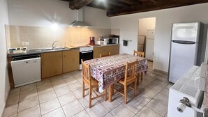 Een kleine keuken met betegelde vloer, houten kastjes, een eettafel met stoelen, een koelkast, een magnetron, een fornuis en een vaatwasser.