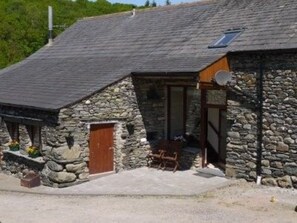 A stone building with a sloped roof, a wooden door, and a small porch area. - Lind End cottage warm, comfortable and inviting, super views (Broughton-in-Furness)