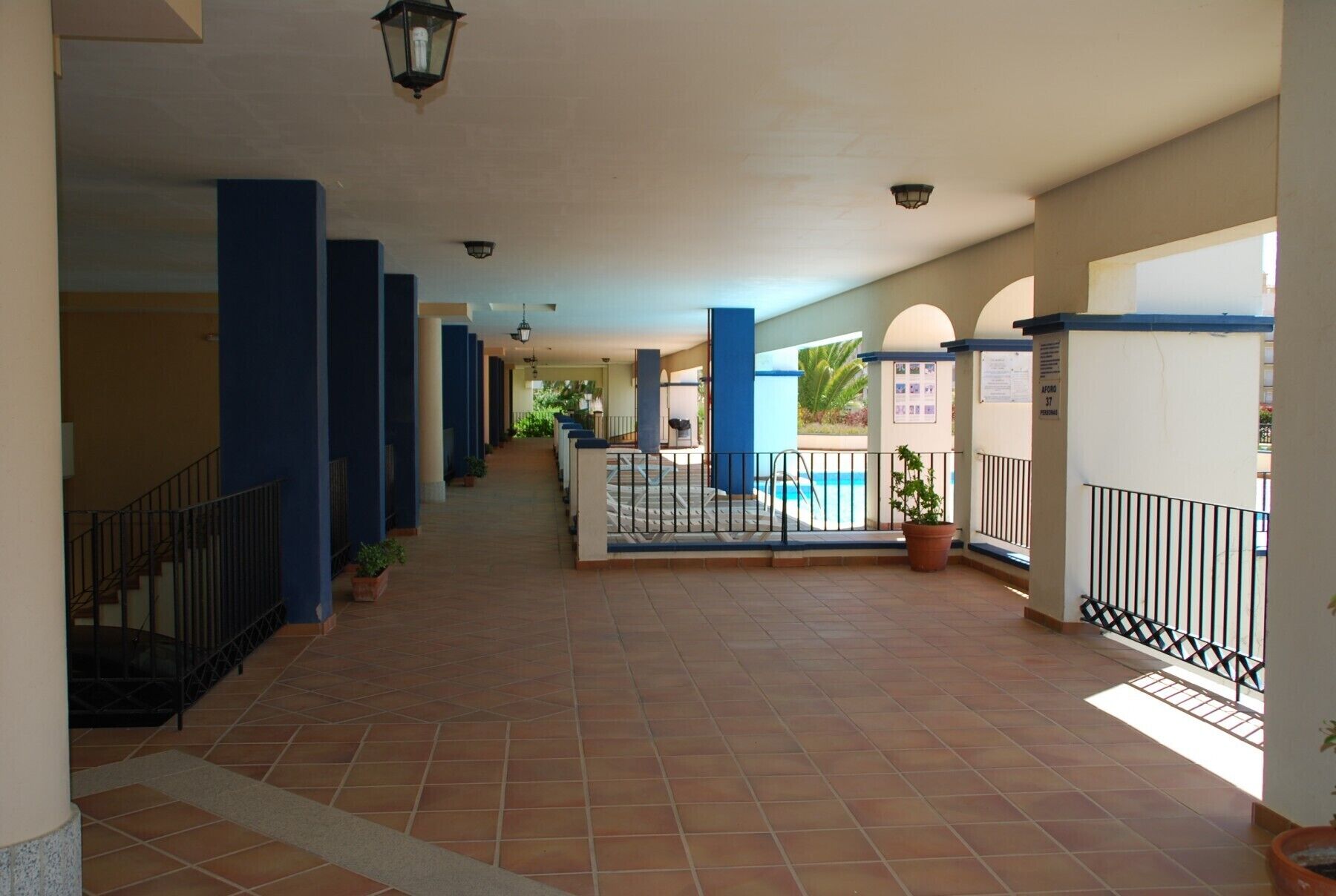 A covered corridor with tiled flooring, blue pillars, and a pool area visible through the glass.
