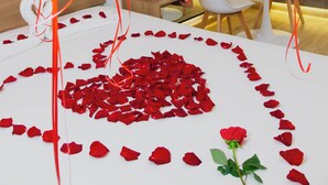 Una habitación con una mesa blanca, pétalos de rosa roja formando un corazón, una sola rosa y globos rojos.
