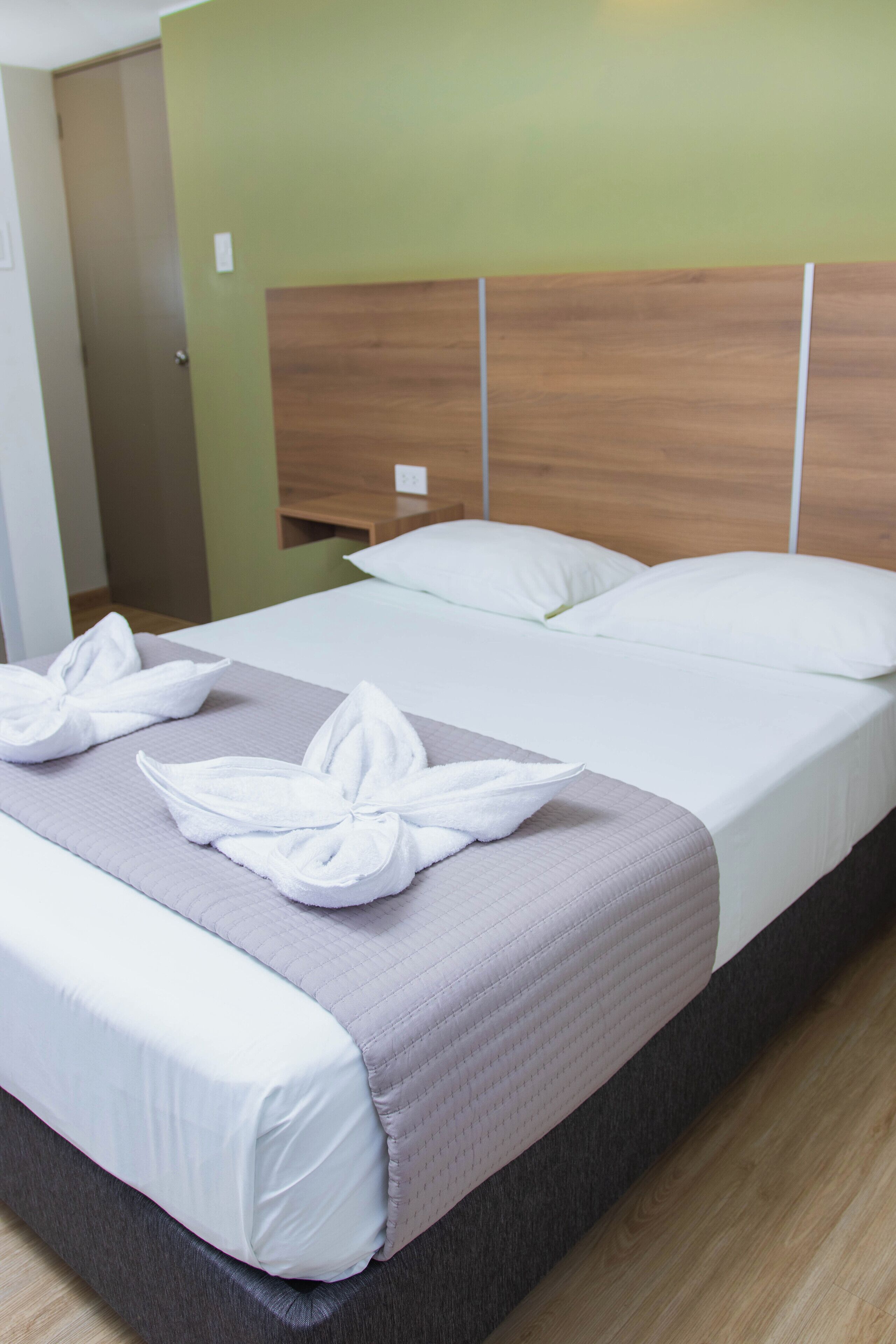 A neatly made bed with white linens and a grey blanket, a wooden headboard, and a green wall.