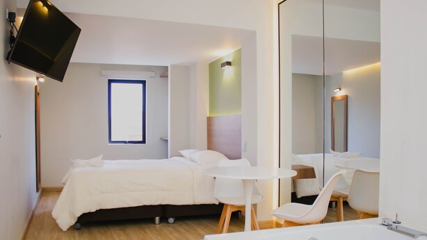 Una habitación de hotel moderna con una cama, una pequeña mesa redonda, una silla, un espejo grande y un televisor en la pared.