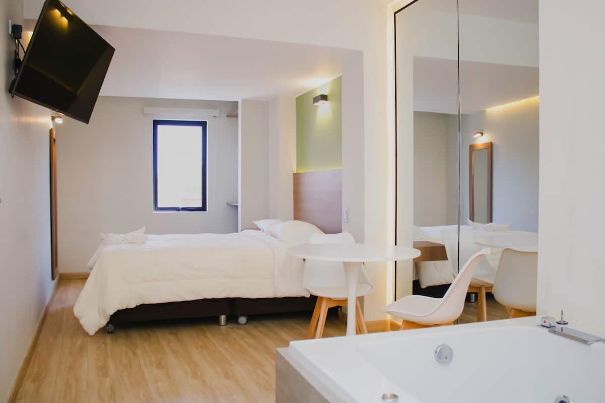 A modern hotel room with a bed, a small round table, a chair, a large mirror, and a TV mounted on the wall.