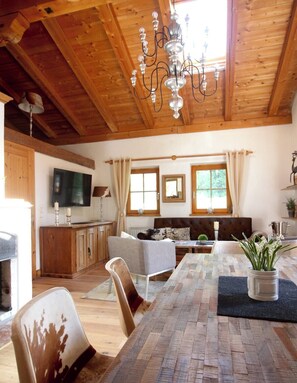 A wooden interior with a dining table, chairs, a sofa, a TV, and a chandelier.