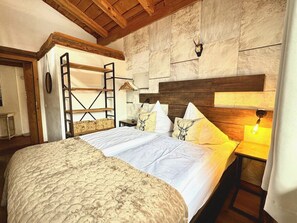 A bedroom with a bed, a wooden ladder, a nightstand, a lamp, and a stone wall.