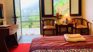 A hotel room with a bed, two chairs, a desk, and a large window with a view of a green landscape.