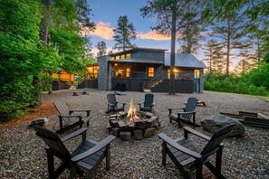 Una cabaña con chimenea de piedra rodeada de sillas Adirondack, ubicada en un área boscosa con vista al edificio.