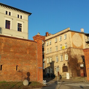 Vista de una calle con un edificio que tiene un reloj en su fachada, un muro de ladrillo y un cielo azul despejado.