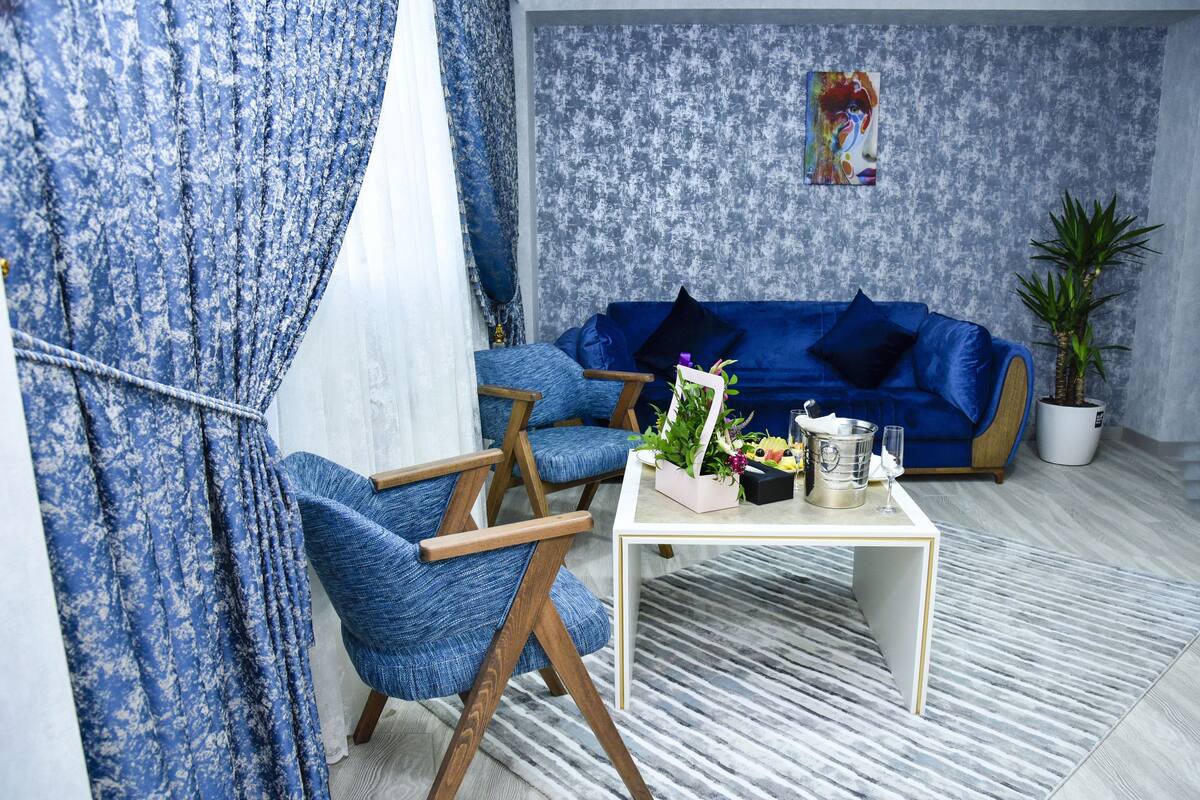 Un salon avec un canapé bleu, deux fauteuils bleus, une table basse blanche ornée d’une plante et d’un vase, et un tableau accroché au mur.