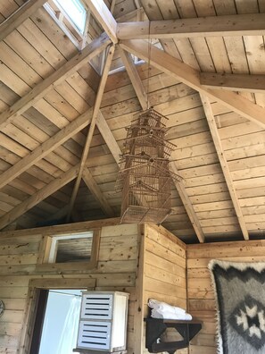 Interior de madera con techo inclinado, una jaula colgante, una ventana con contraventanas y un estante en la pared con toallas.