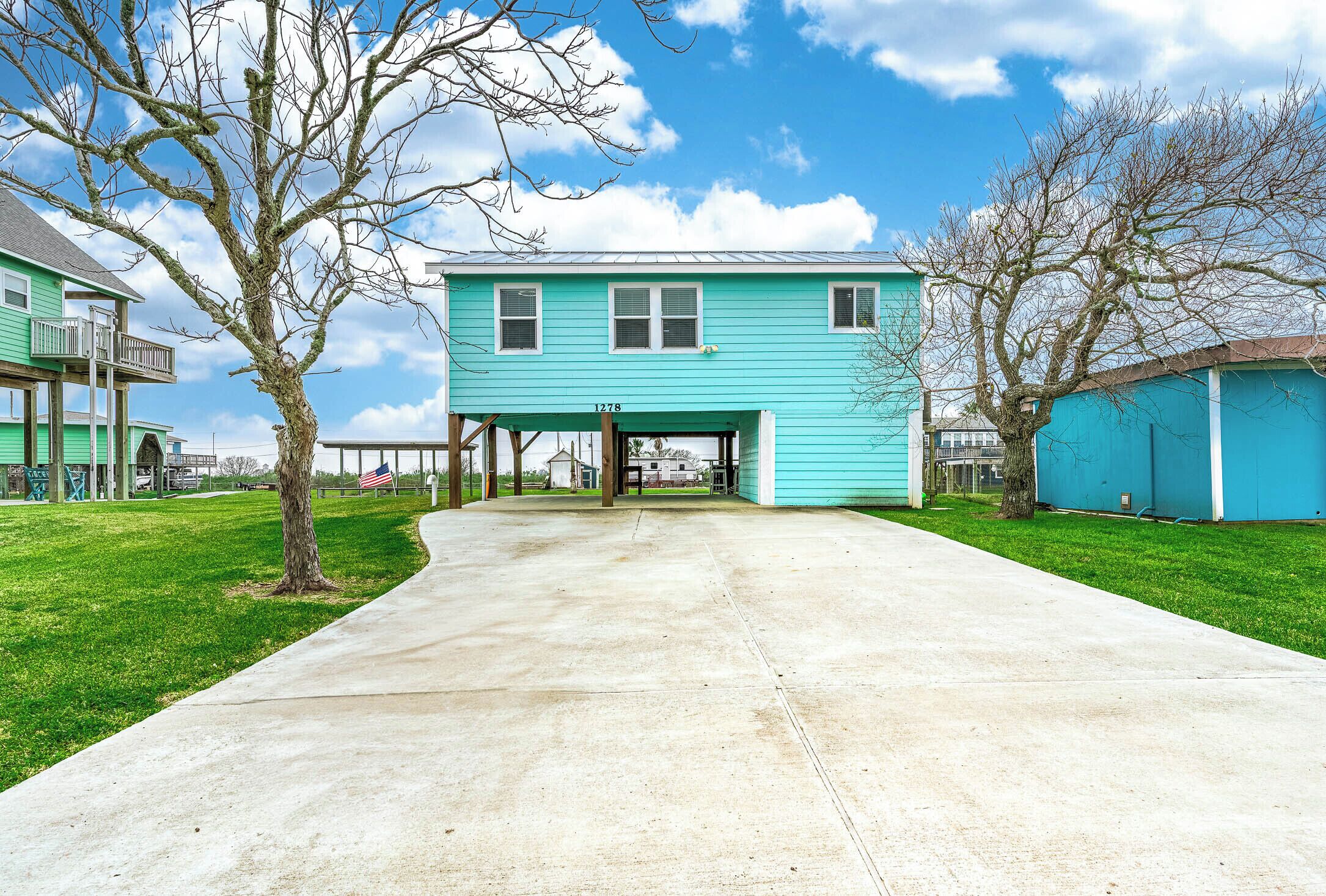 Une maison turquoise avec une allée couverte, entourée de pelouses verdoyantes et d’arbres.
