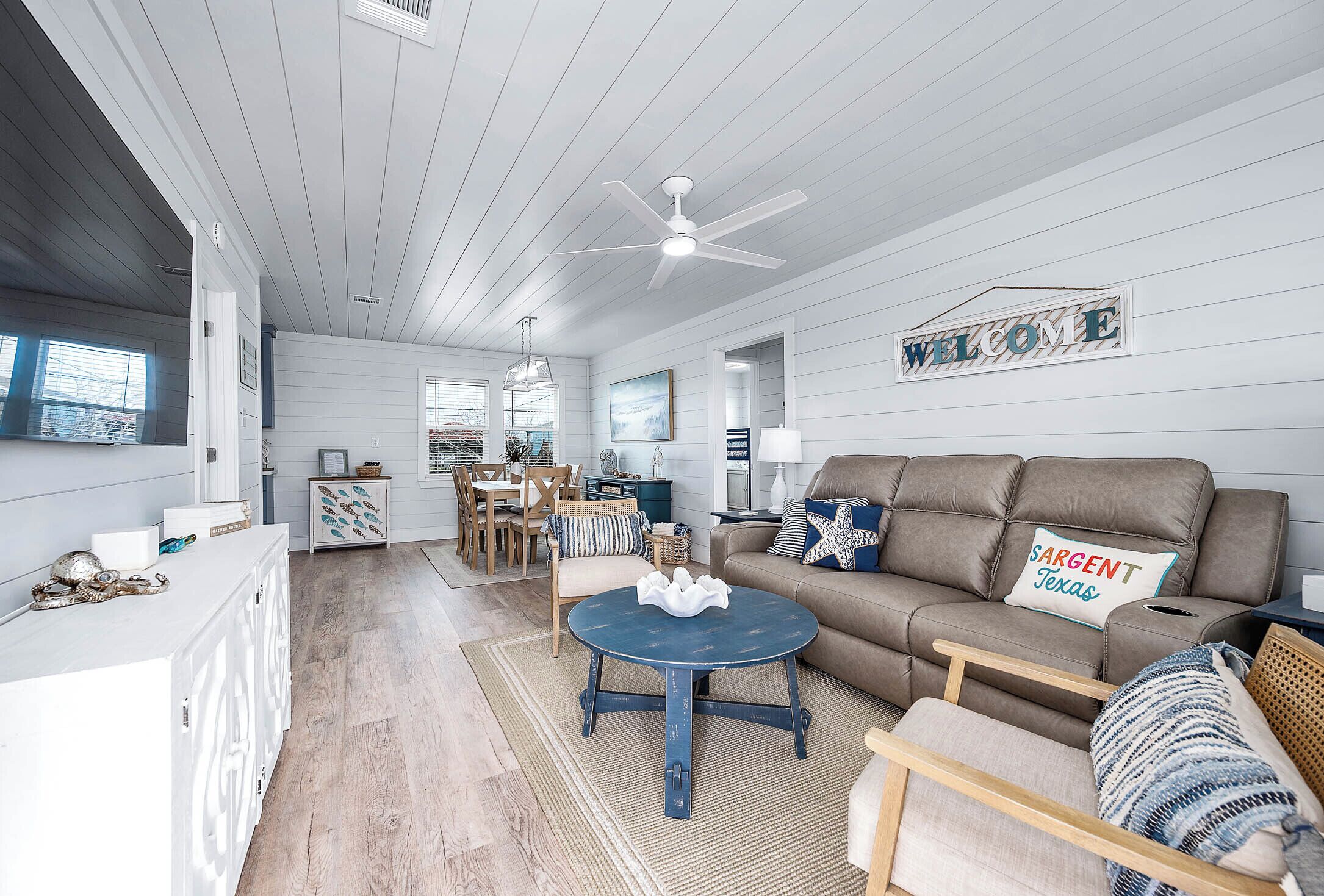A spacious living room with a blue coffee table, a brown sofa with a 'SARGENT Texas' pillow, and a 'WELCOME' sign on the wall.