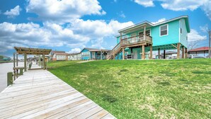 A waterfront property with a wooden dock, a grassy lawn, and a turquoise house with a balcony.