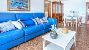 A living room with a blue sofa, a white coffee table, and a painting of a harbor scene. - Lovely apartment for 6 people with WIFI and TV (L'escala)