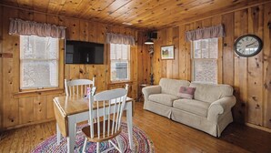 Una habitación con paneles de madera, un televisor, un sofá, una mesa de comedor con sillas y un reloj en la pared.