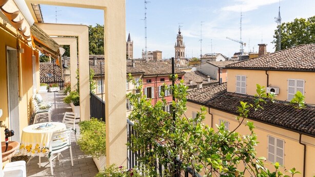 Un balcon avec une table et des chaises, donnant sur une ville historique avec des bâtiments traditionnels et un imposant beffroi.