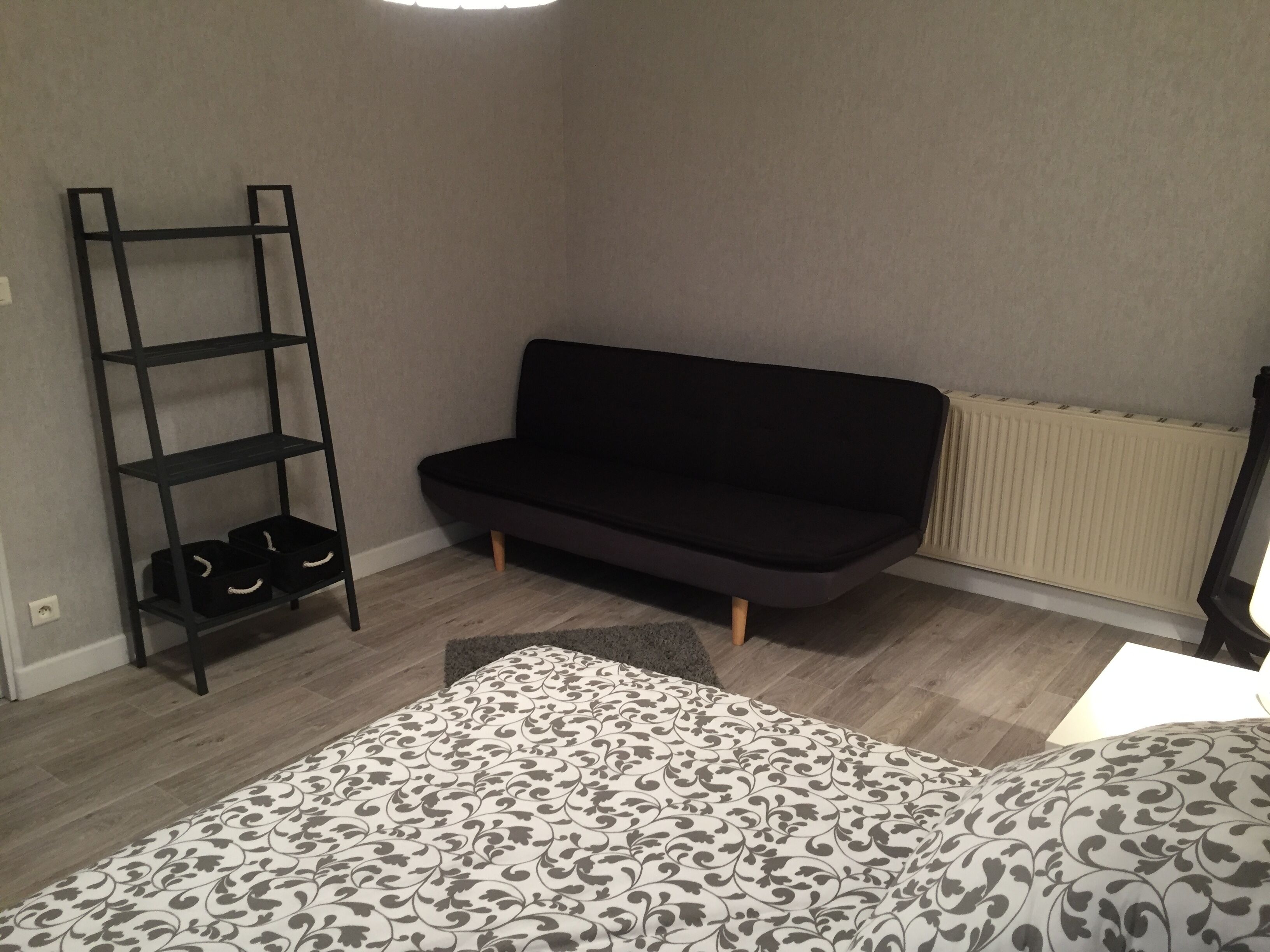Une pièce avec un canapé noir, un plancher en bois, une couvre-lit à motifs et une étagère en métal noir où sont rangées des chaussures.