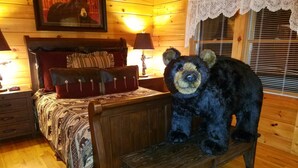 A wooden cabin interior with a bed, a nightstand, a lamp, a window with blinds, and a bear statue on a bench.