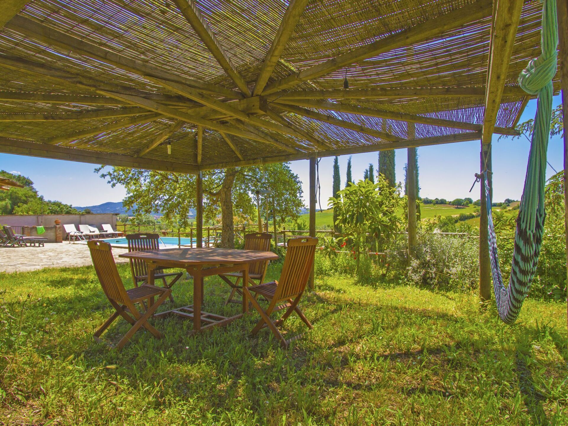 Een terras buiten met een houten tafel en stoelen, een zwembad op de achtergrond en uitzicht op het landschap.