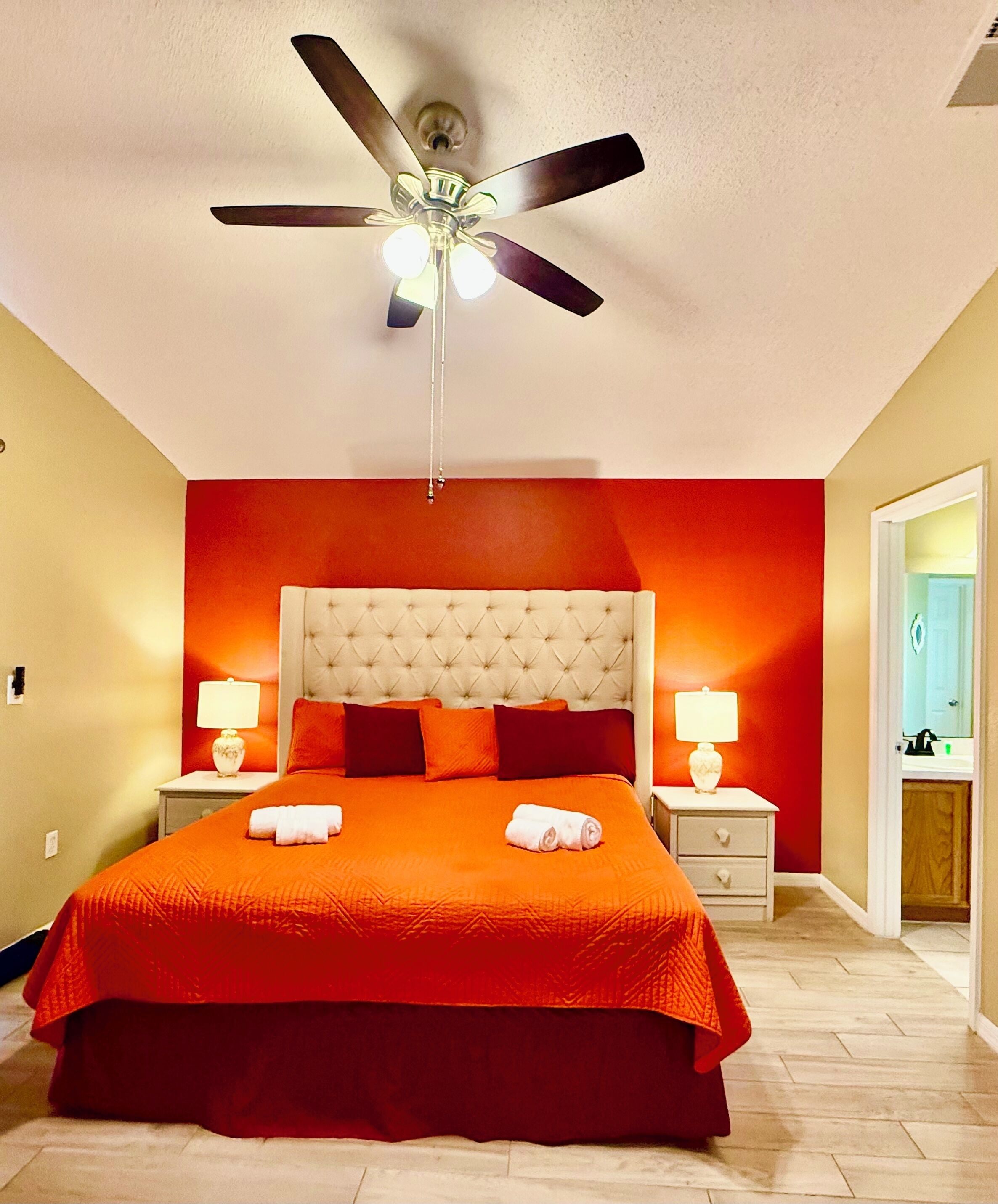 A hotel room with a large bed, a ceiling fan, two bedside tables with lamps, and a bathroom visible through an open door.