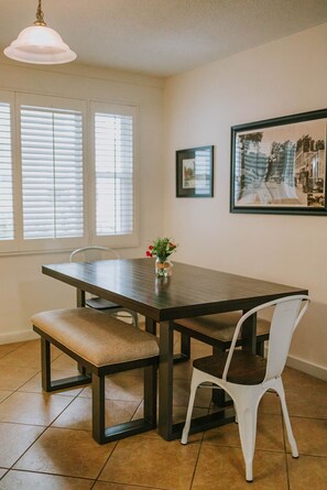 Un comedor con una mesa de madera oscura, dos bancos acolchados, una silla blanca, una lámpara colgante y un cuadro en la pared.