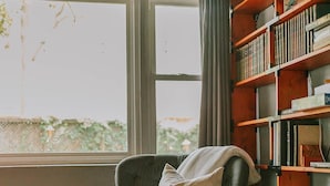 A room with a large window, a grey armchair with a cushion, a small table, and a wooden bookshelf filled with books. - Walk to the BEACH and close to DOWNTOWN - the perfect location! The Main Suite (Ocean Springs)