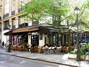 An outdoor cafe with tables and chairs, a tree, and a street in the background.