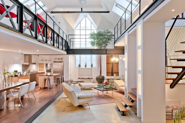 A spacious loft-style interior with a mezzanine, open kitchen, and a large window providing natural light.