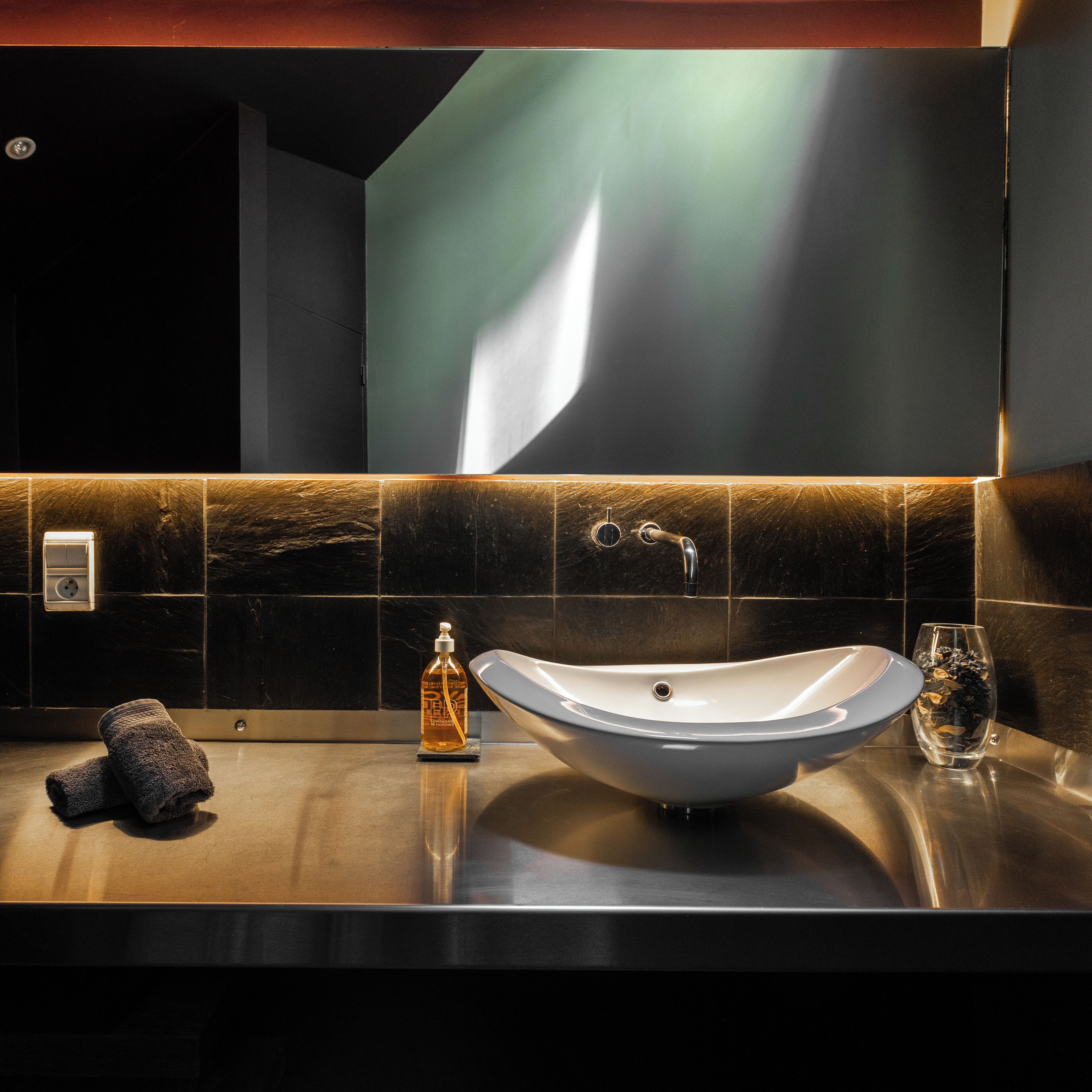 Une salle de bain moderne avec un grand lavabo sur pied, des carreaux foncés et un miroir fixé au mur.