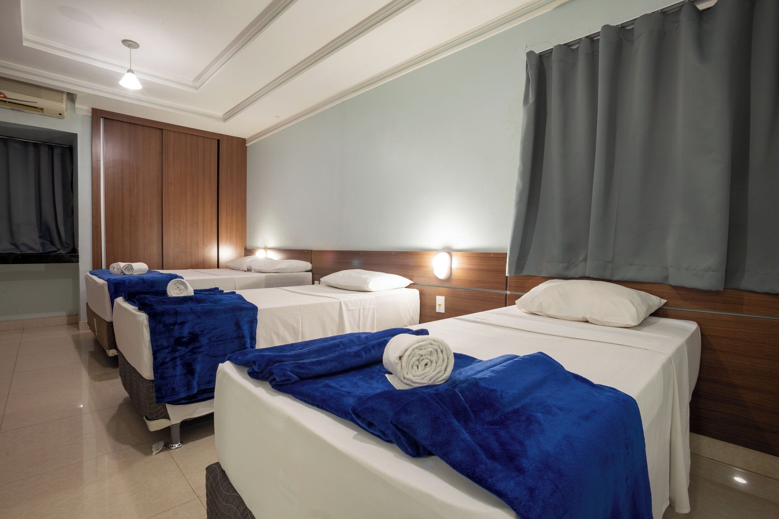 A hotel room with three beds, each with a white mattress and blue blankets. There are wooden headboards and a large window with curtains.