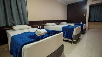 Habitación de hotel con tres camas, cada una con una manta azul y almohadas blancas. El cuarto tiene piso de baldosas y un armario grande.