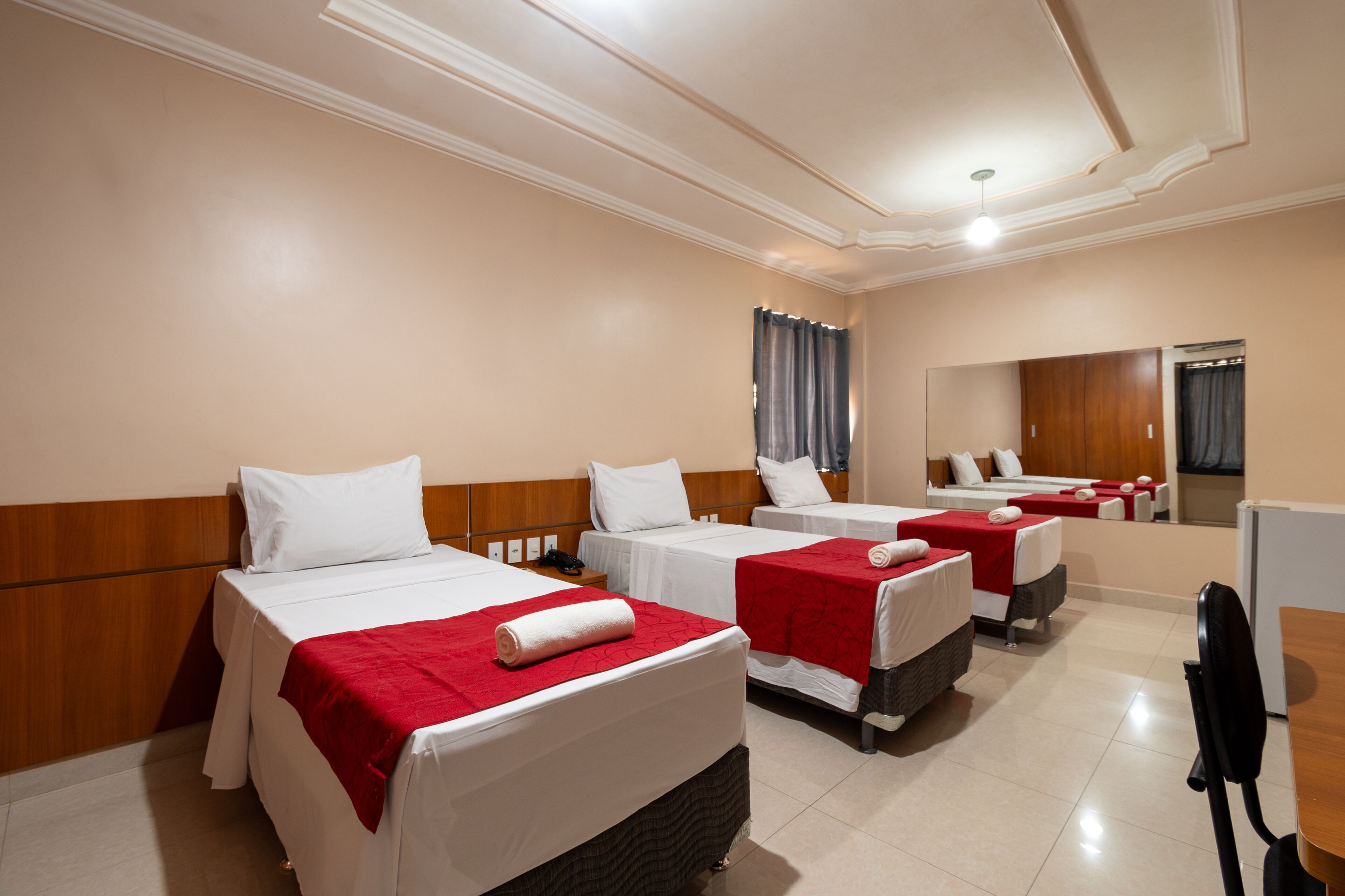 A hotel room with three single beds, each with a red blanket and white pillows. The room has a wooden headboard, a large mirror, and a wooden desk with a chair.