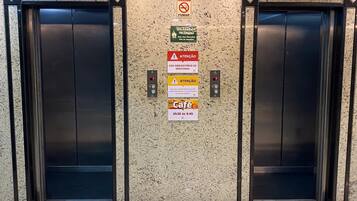 Un ascensor con múltiples señales, incluyendo una señal de no fumar, una señal de emergencia por incendio, una señal obligatoria de mascarilla, una señal de precaución y una señal con los horarios de atención de una cafetería.
